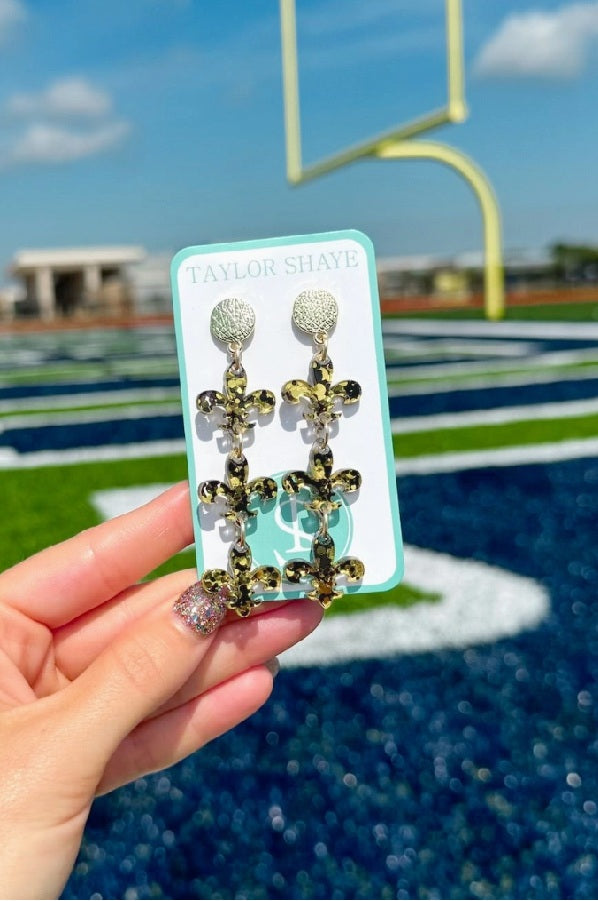Confetti Fleur De Lis Earrings by Taylor Shaye in Black/Gold on card at a football field